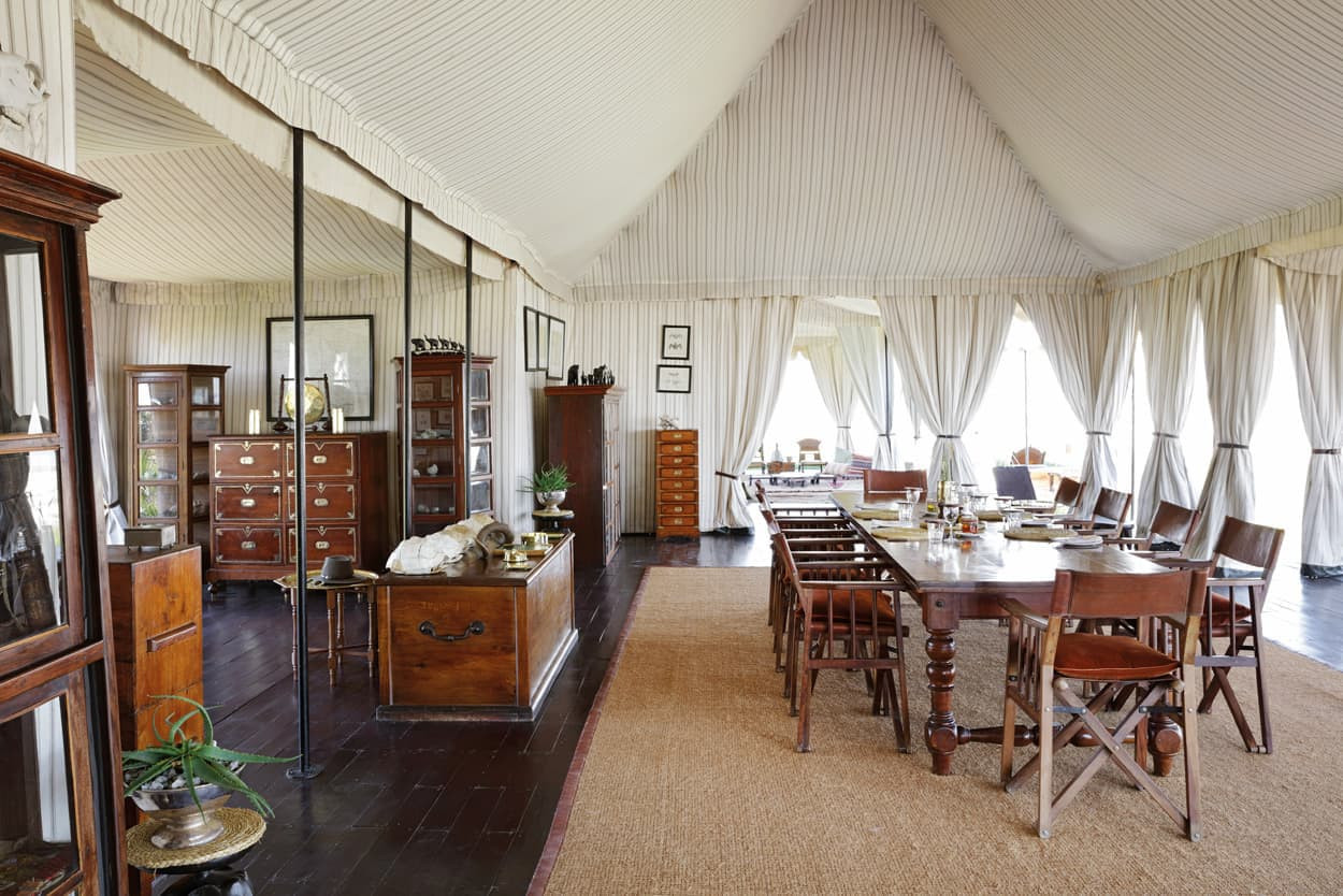 A spacious, elegantly designed interior featuring a dining area with a long wooden table surrounded by chairs. The room is filled with vintage furnishings, including chests of drawers and display cabinets. Large windows draped with light fabric allow natural light to fill the space, accentuating the soft color palette and textures of the decor.