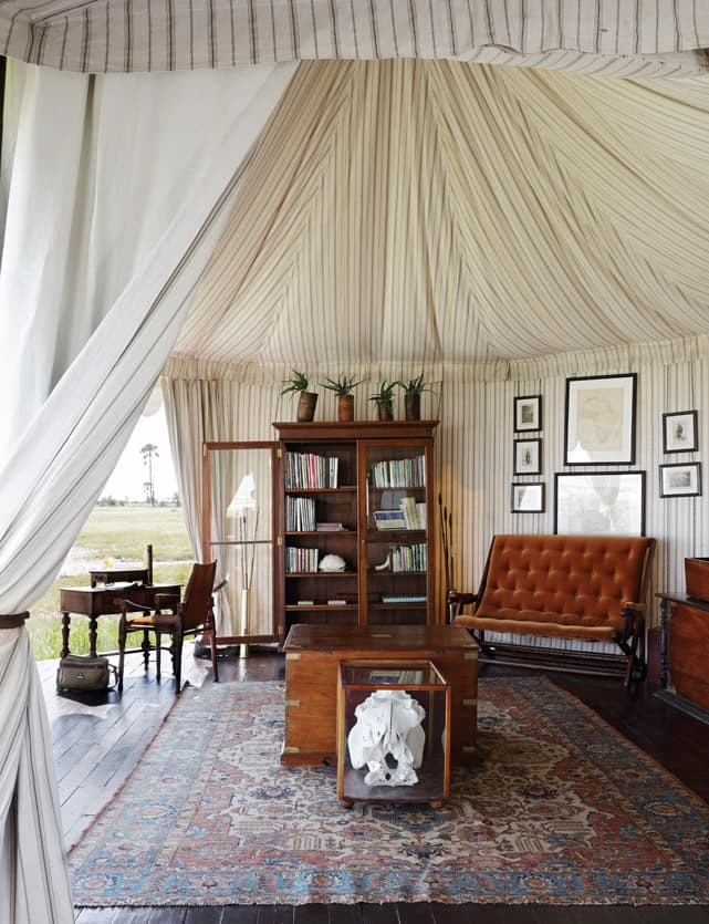 A cozy, well-decorated tent interior featuring a vintage wooden bookshelf filled with books, a stylish brown tufted sofa, a low wooden coffee table with a decorative item, and framed artwork on the walls. Soft, draped fabric lines the ceiling and walls, while a patterned rug adorns the floor. A small table is set in one corner, adding to the inviting atmosphere.