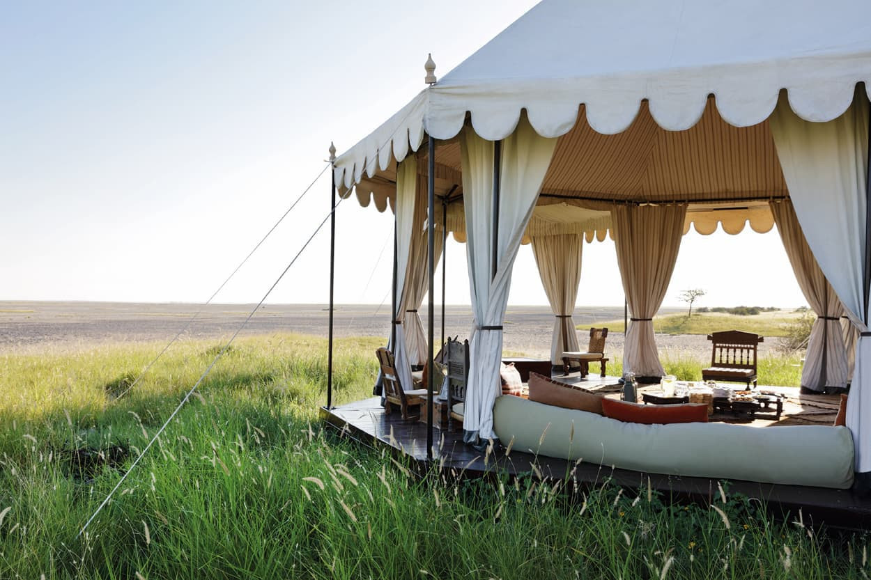 A luxurious tented lodge in a grassy landscape, featuring elegant seating and draped fabric. The tent is open to the outdoors, showcasing a scenic view of the surrounding plains.