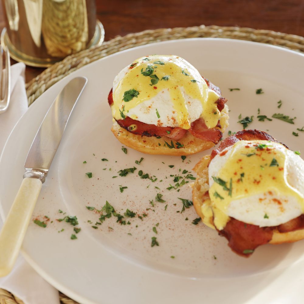 Two eggs benedict on toasted English muffins, topped with poached eggs, crispy bacon, and drizzled with hollandaise sauce, garnished with parsley and a sprinkle of paprika.