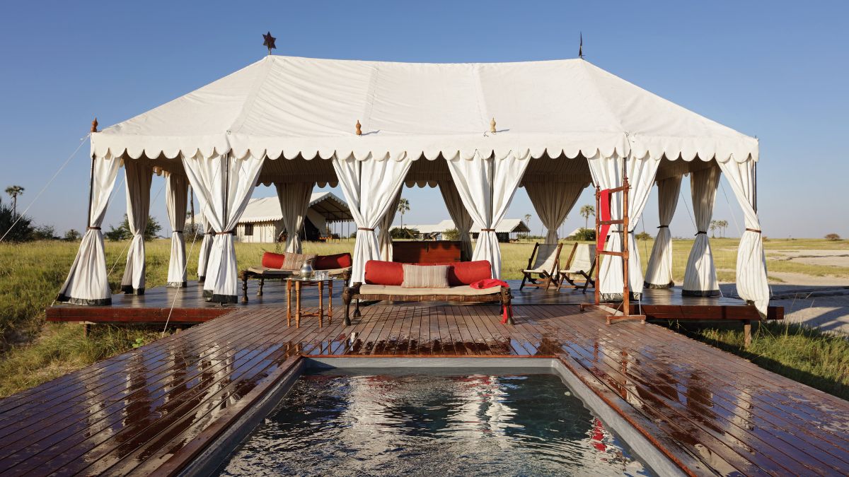 A luxurious tent on a wooden deck in a serene outdoor setting, featuring a small private pool and stylish seating with red cushions. The sky is clear and blue, with distant palm trees visible.