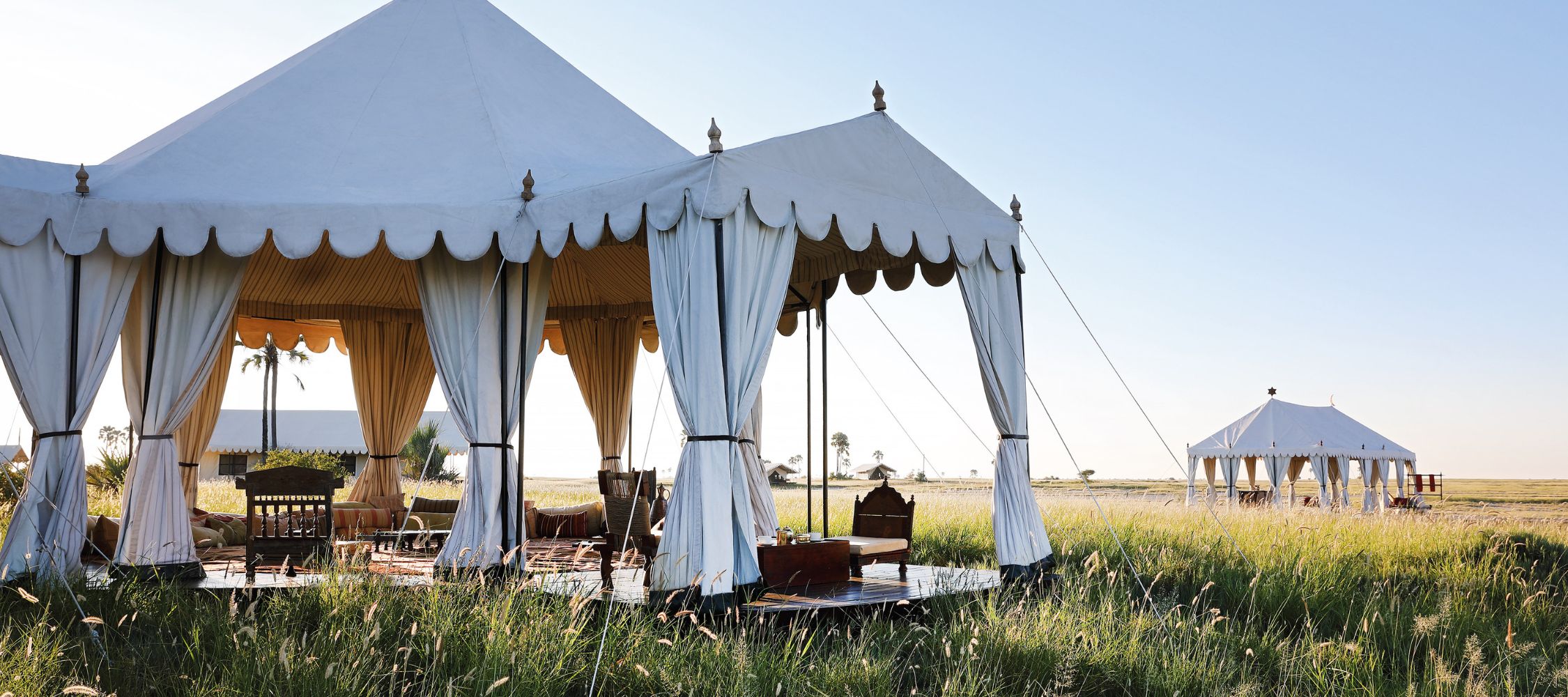 Luxury safari tents set against a clear blue sky, surrounded by lush green grass. The tents feature elegant drapery and wooden furniture on a wooden deck. A second, smaller tent is visible in the background.