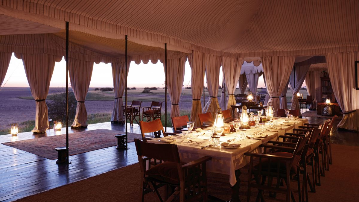 Elegant dining area inside a spacious tent, featuring a long table set with fine china and glassware. Soft lighting from lanterns creates a warm ambiance, while large open windows provide views of the serene outdoor landscape at dusk.