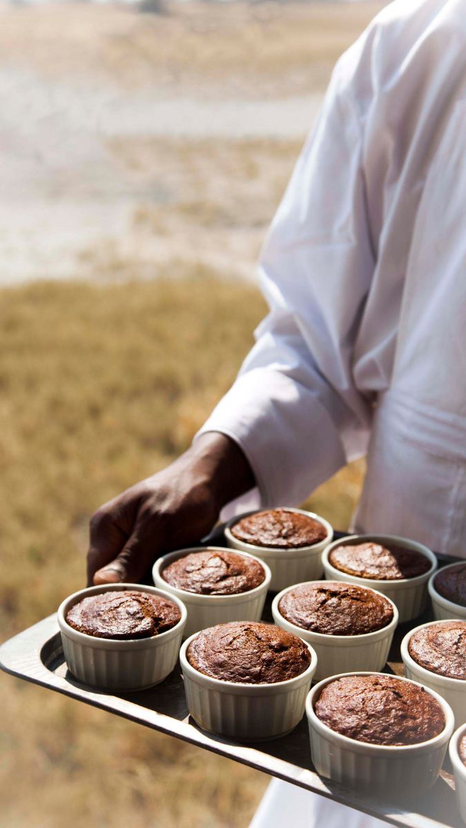 A person holding a tray with several small, round chocolate desserts in white ramekins, set against a blurred outdoor background with grass.