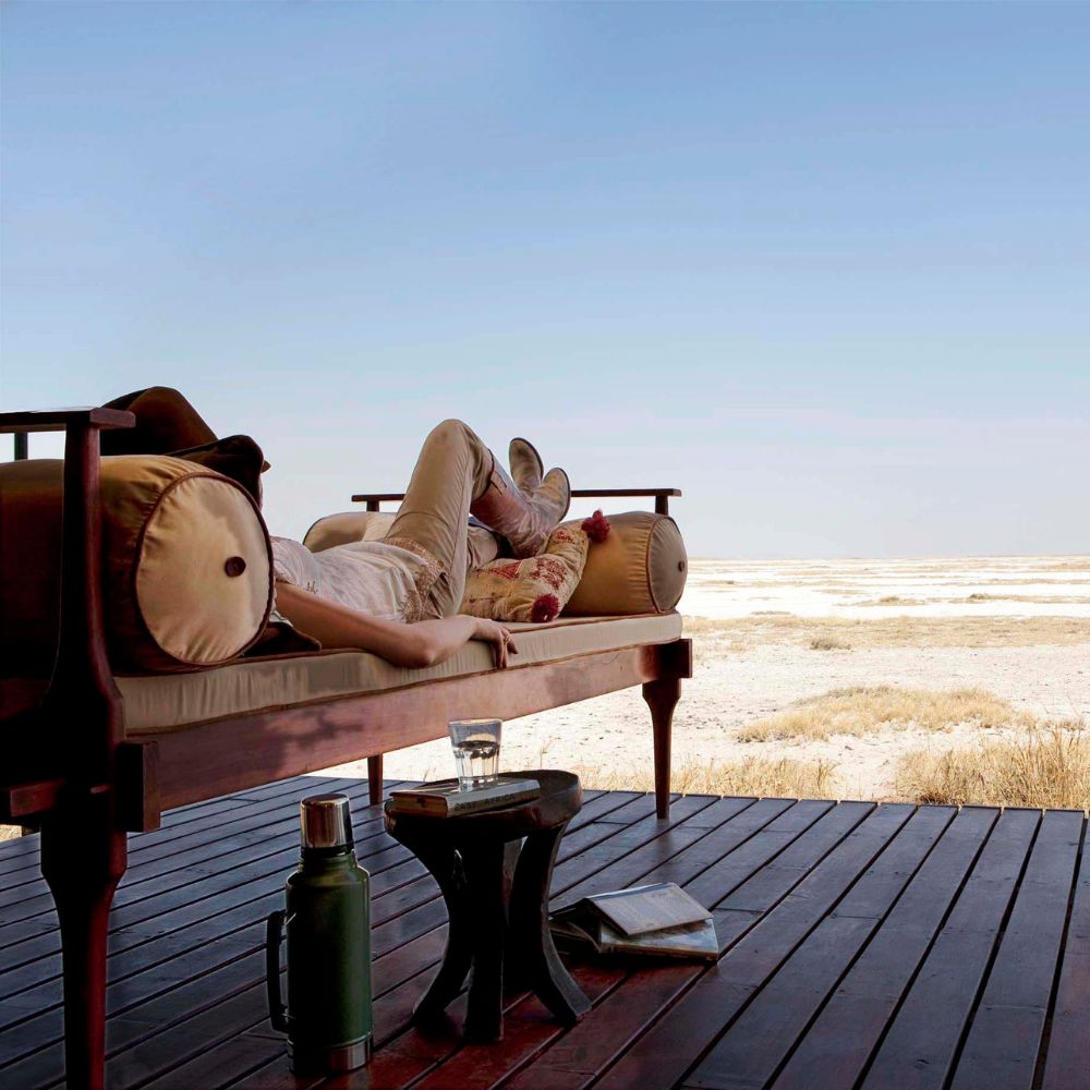 A person lies on a wooden daybed on a deck, facing a vast, open landscape under a clear blue sky. A small table beside them holds a glass and a book.