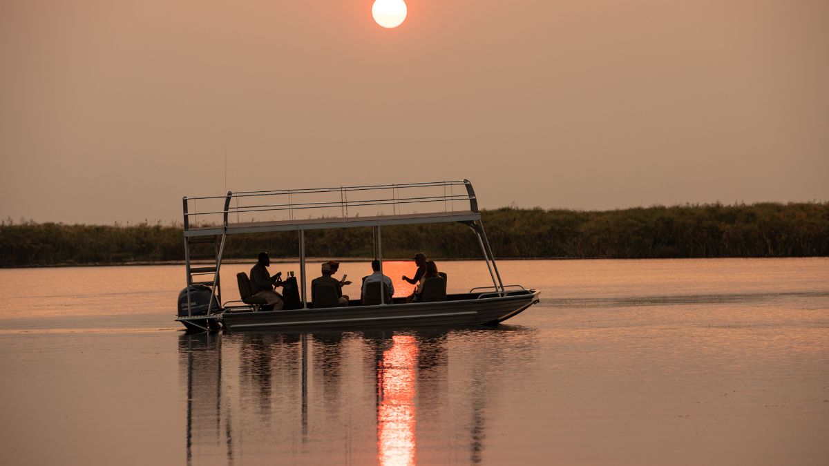 A boat with four people floats on calm water at sunset. The sun sets low on the horizon, casting a golden glow over the scene. Silhouetted figures enjoy the tranquil moment.