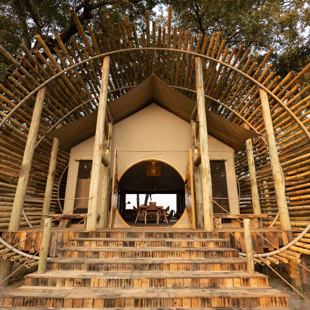 A stylish glamping tent with a wooden entrance and circular bamboo design, set among trees. Steps lead up to the tent, which features large openings and a cozy interior visible through the entrance.