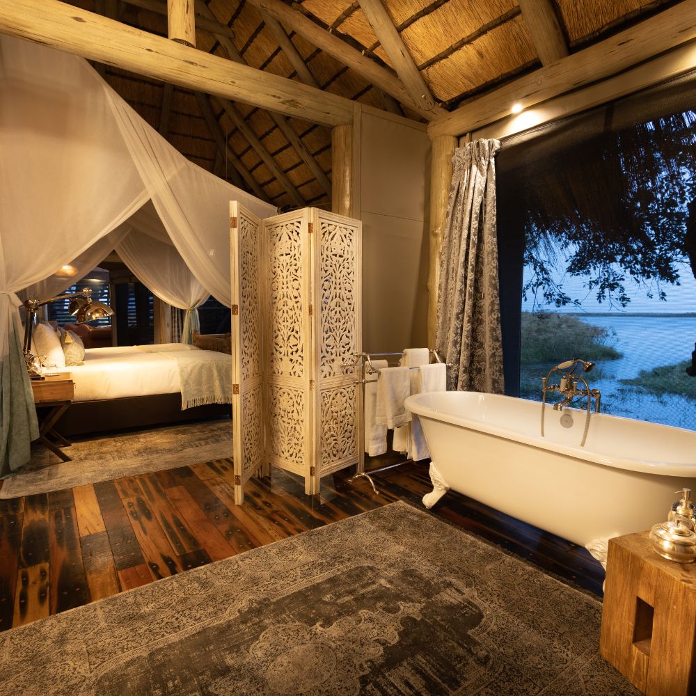 A luxurious bathroom featuring a freestanding bathtub, wooden floors, and a decorative privacy screen. Natural light streams in through large windows, overlooking a serene outdoor view. The room is adorned with rustic wooden beams and a thatched roof, creating a cozy atmosphere.