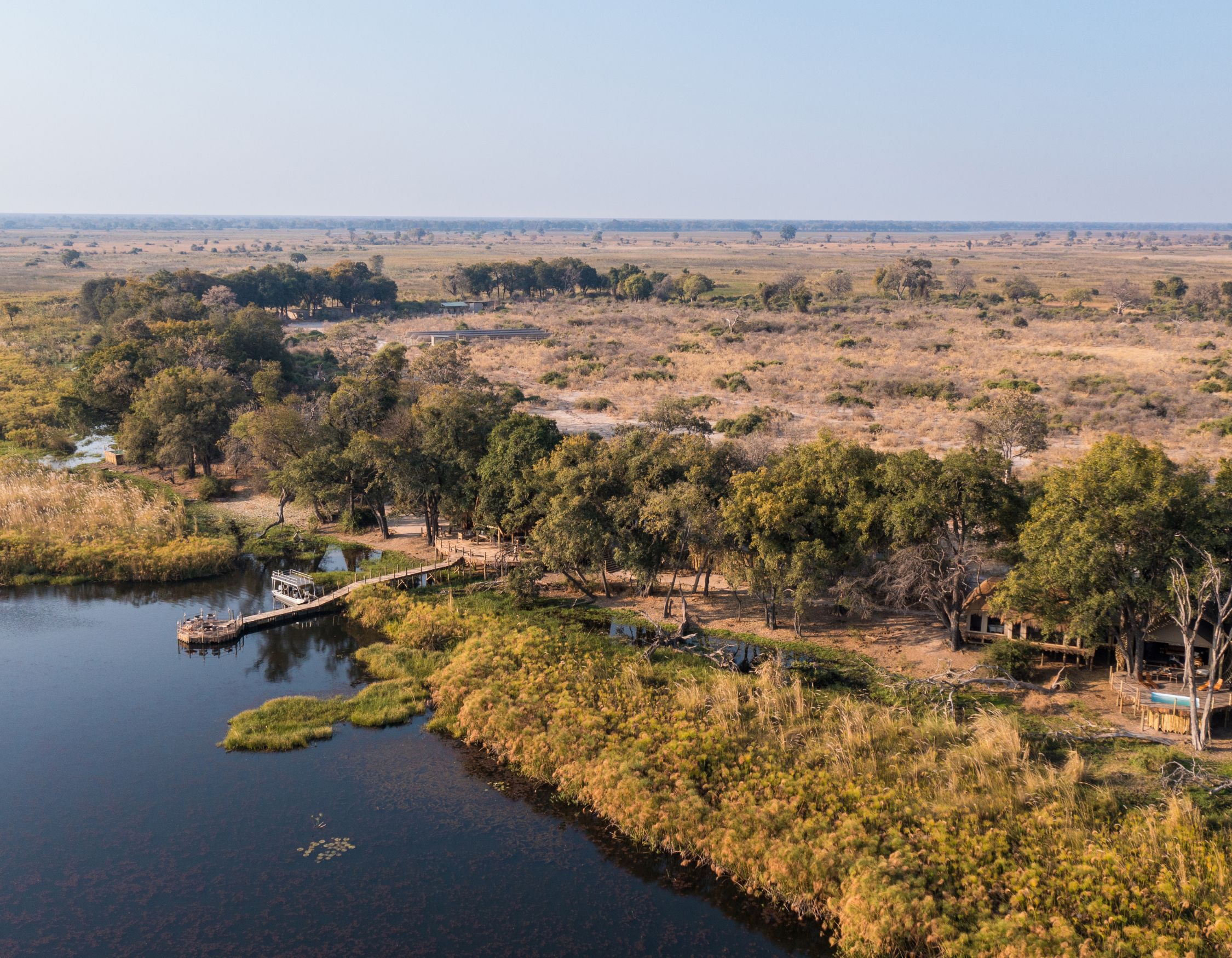Aerial view of a serene riverbank surrounded by lush greenery and sparse grassland, with a wooden dock extending into the water. The landscape features scattered trees and open fields under a clear blue sky.