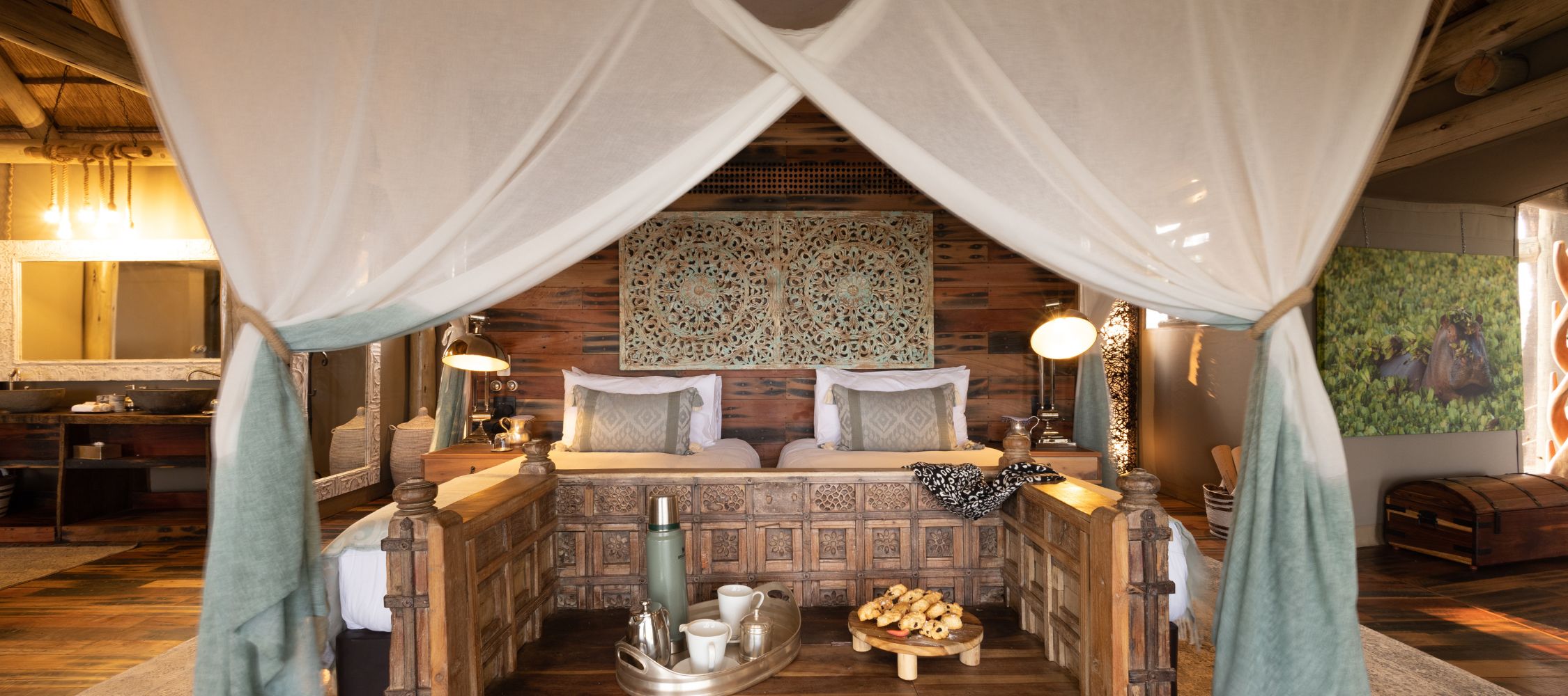 A beautifully designed hotel room featuring a four-poster bed with sheer drapes, intricately carved wooden details, and elegant bedding. The space includes a wooden side table with a plate of pastries and a beverage tray, while soft lighting adds warmth. A decorative wall art piece enhances the rustic ambiance.