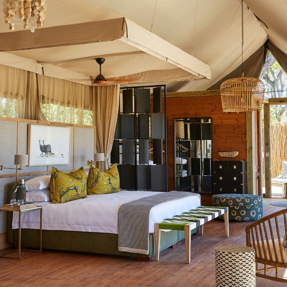 A stylish tented bedroom featuring a large bed with decorative pillows, a patterned bench at the foot, wooden flooring, and woven decor elements. Natural light flows in through the open windows, creating a warm and inviting atmosphere.