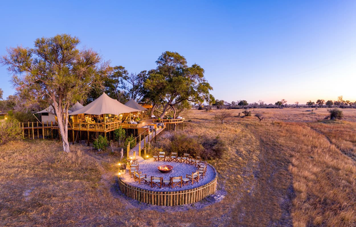 A scenic view of a safari lodge surrounded by tall trees, with a wooden deck and cozy seating around a fire pit. The landscape features golden grasslands under a twilight sky.