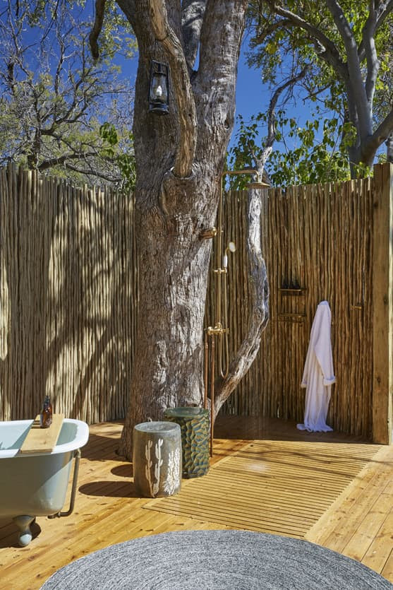 Outdoor bathroom setup featuring a vintage bathtub beside a wooden deck, surrounded by a rustic bamboo fence and a large tree. A white robe hangs on a hook, with outdoor shower fixtures attached to the tree. Bright blue sky and green foliage in the background.