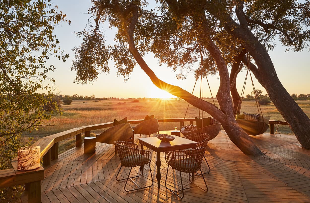 A picturesque outdoor deck featuring wooden flooring, surrounded by trees, with a table and chairs set up. Sunlight streams through the branches as the sun sets over a grassy landscape, creating a tranquil ambiance.