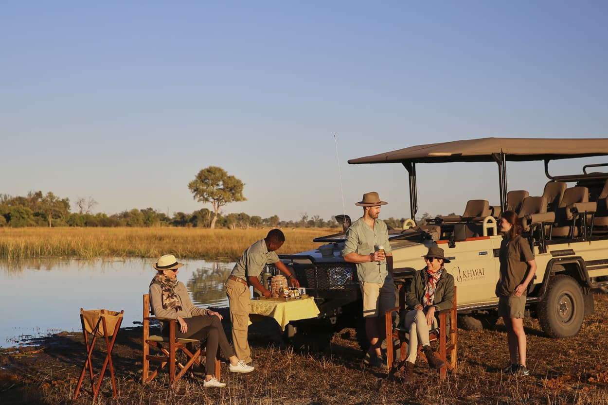 A group of four people enjoying a safari picnic near a river, with a safari vehicle parked in the background. One person is serving food at a table, while others relax in folding chairs. The setting is serene, with grasslands and a clear blue sky.