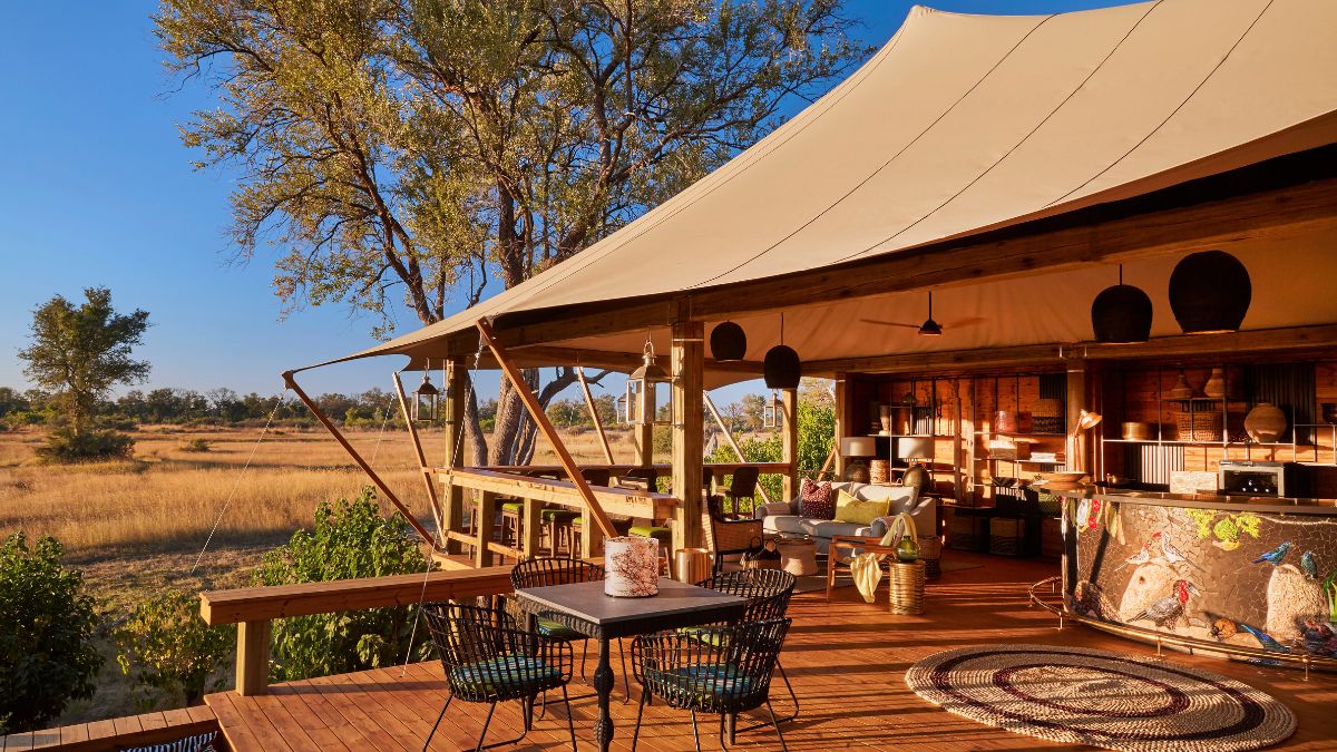 A luxurious tented lodge surrounded by greenery and open savanna. The spacious deck features stylish seating, a dining area, and a vibrant bar. Trees provide shade under a clear blue sky.