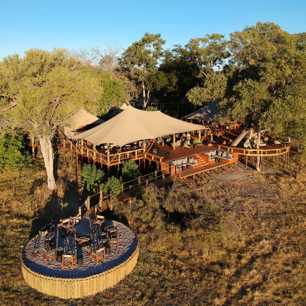 A spacious outdoor dining area with a large, tan canopy surrounded by trees. Wooden decks and tables are set up for guests. The scene is bathed in warm sunlight, highlighting the natural surroundings.
