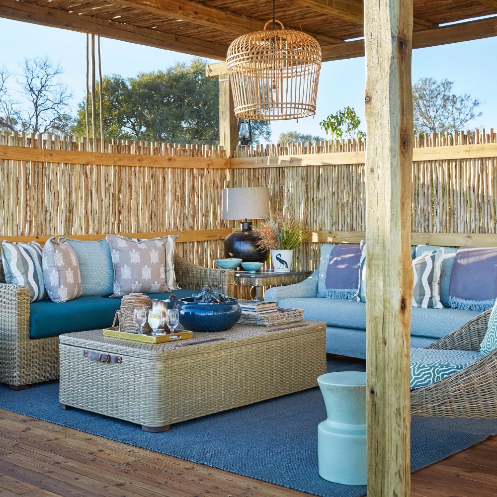 A stylish outdoor seating area featuring wicker furniture with blue cushions, a coffee table topped with decorative items, and a hanging pendant light. The space is surrounded by a wooden fence and has a natural wood floor. Decorative pillows and a rug add comfort and style.