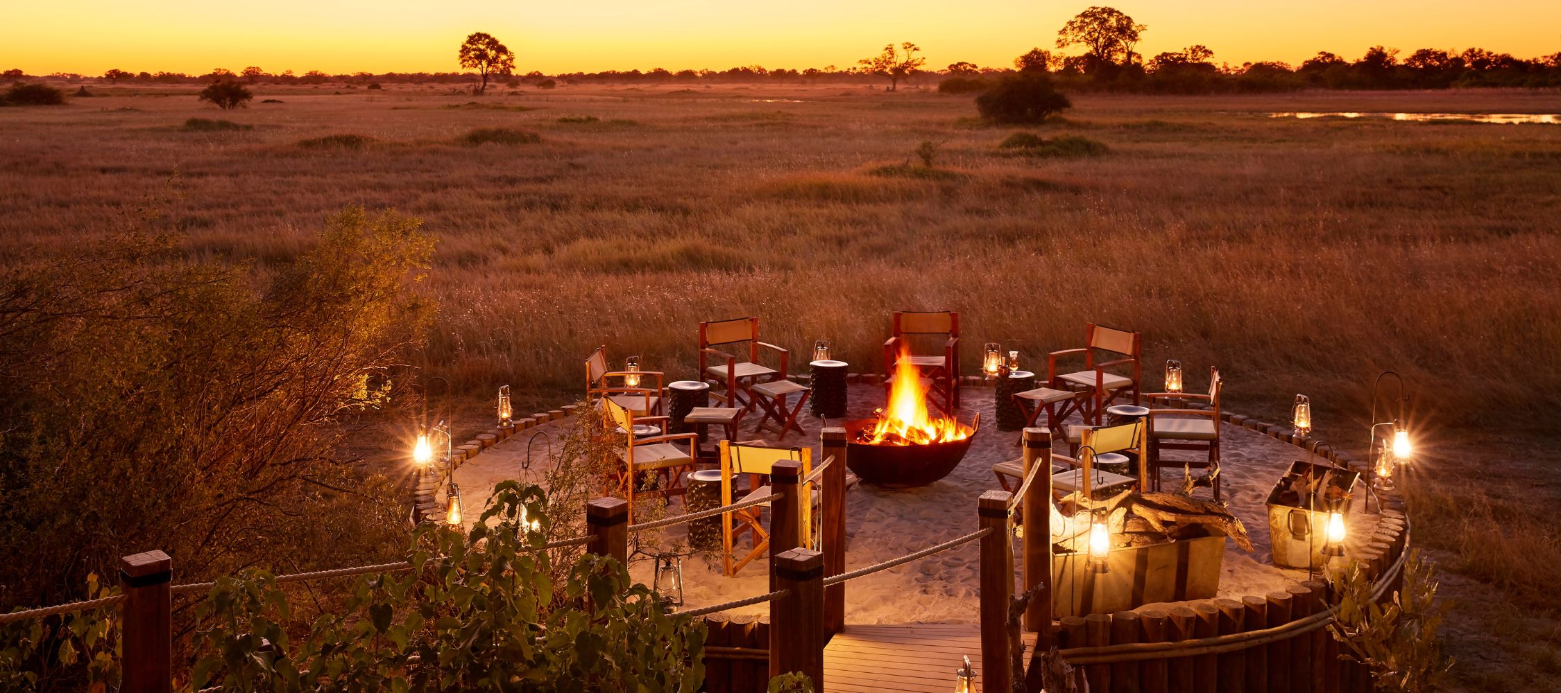 A cozy campfire setup surrounded by chairs, illuminated by lanterns, against a backdrop of a vast savannah at sunset. The warm glow of the fire contrasts with the dusky orange sky and distant trees.