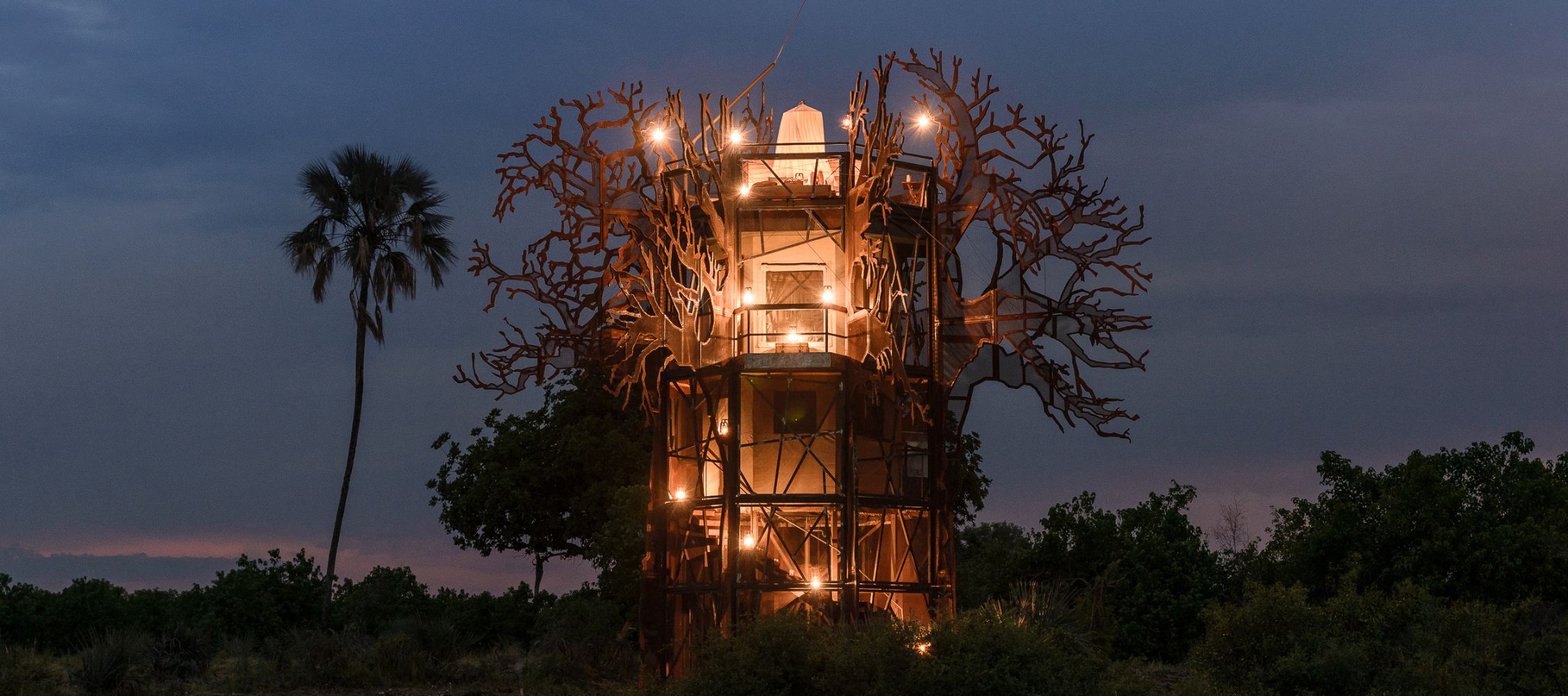 A tall, modern treehouse structure resembling a tree with sprawling branches, illuminated by warm lights, standing in a natural setting at dusk.