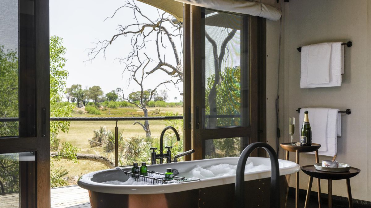 A luxurious bathroom featuring a freestanding bathtub filled with bubbles. Large windows provide a view of a serene outdoor landscape with trees and grass. Two glasses of champagne sit beside the tub, accompanied by a bottle on a small side table. Clean white towels hang neatly on the wall.