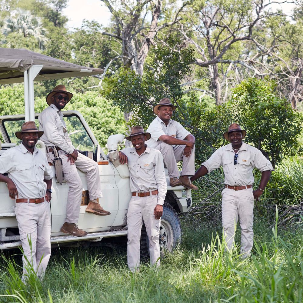 Five safari guides stand beside a white jeep in a lush green landscape. They are wearing light-colored shirts and hats, smiling and posing confidently. Trees and foliage create a natural backdrop.