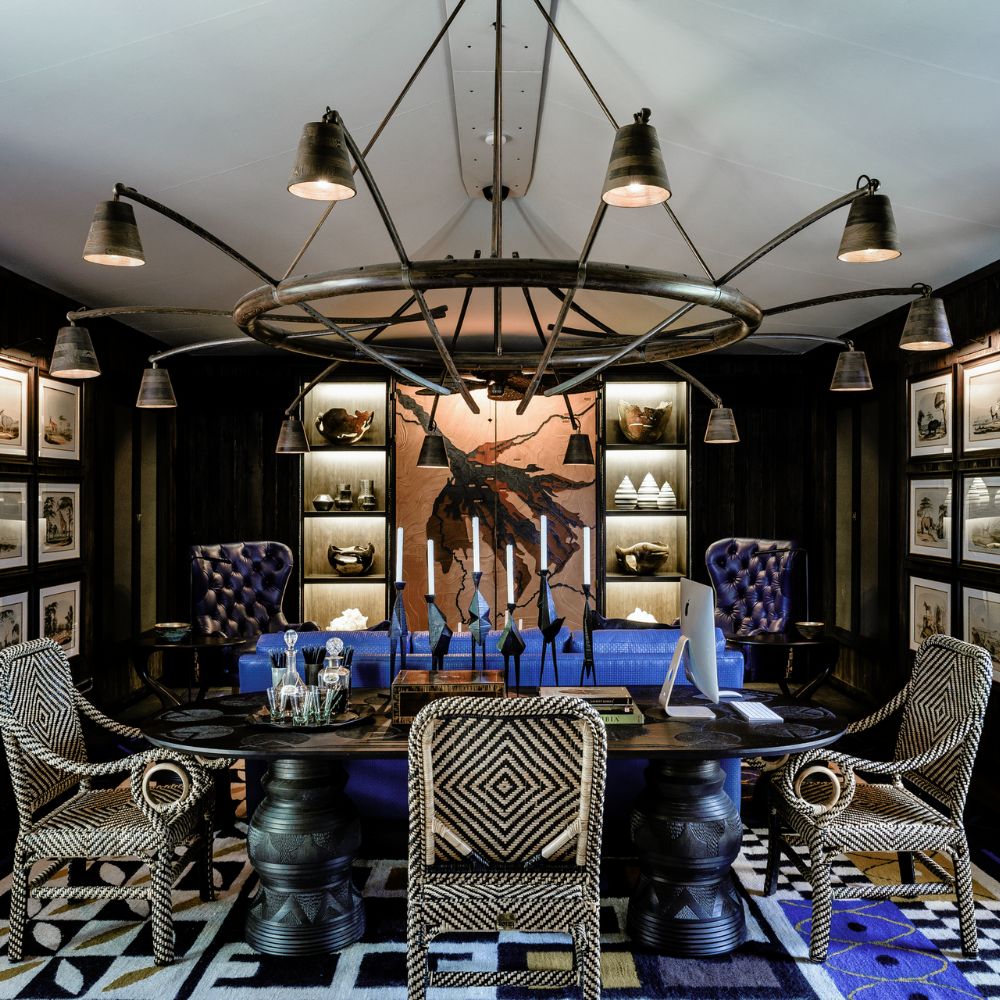 A stylish office featuring a large circular chandelier, dark wooden furniture, and ornate chairs. The walls are decorated with framed black and white photographs and a central piece of art. A blue sofa adds a pop of colour, complemented by a geometric patterned rug.