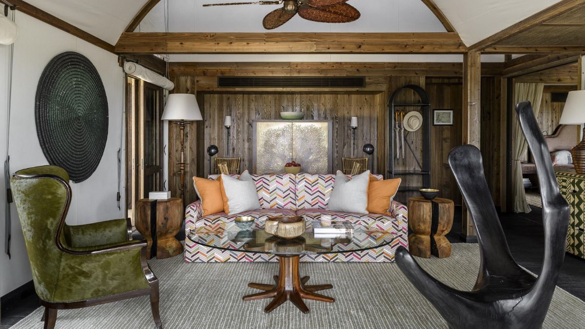 A stylish living area featuring a colorful patterned sofa with orange and cream pillows, two unique chairs, a round wooden coffee table, and wooden walls. The space is bright and inviting, with decorative elements and a ceiling fan.