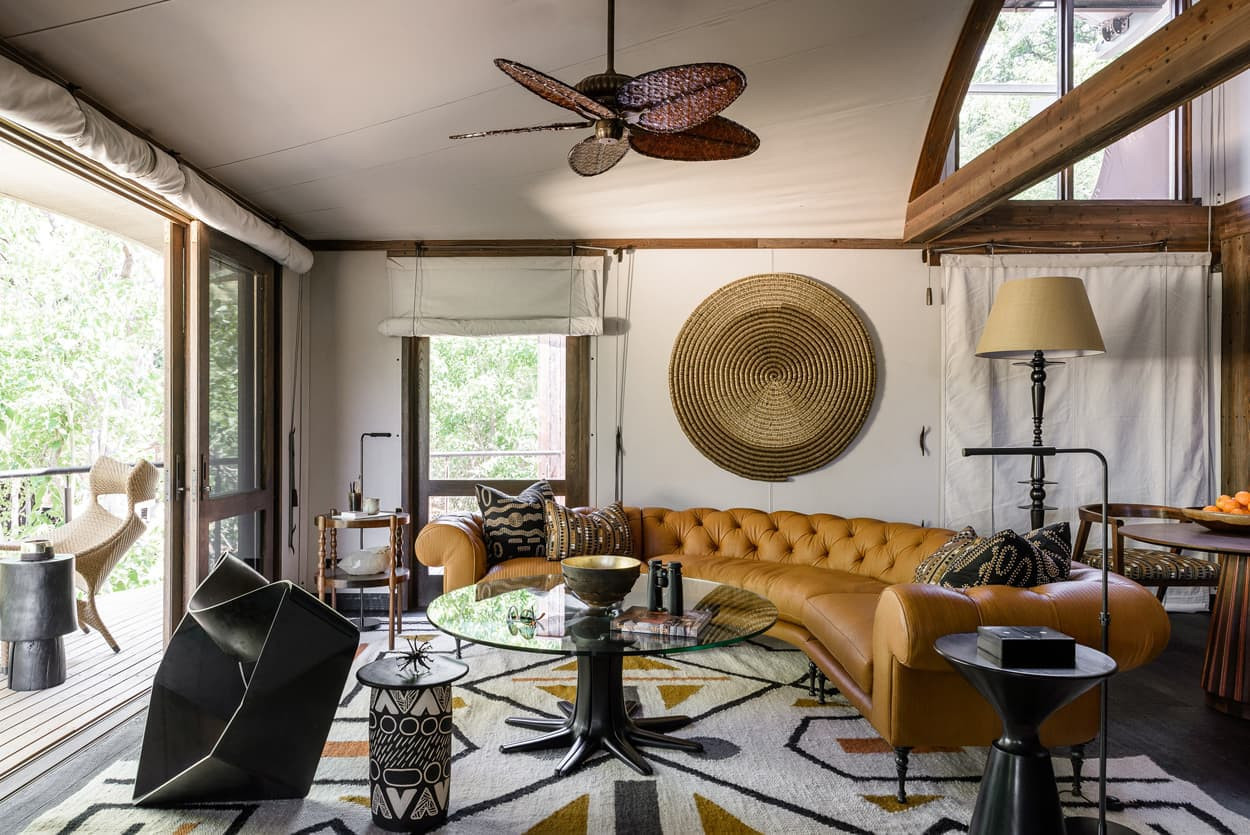 A stylish living room features a tan leather chesterfield sofa, a round glass coffee table, and geometric decor. Natural light streams in through large windows, highlighting a woven wall hanging and a patterned area rug. A ceiling fan with wooden blades adds a touch of elegance.