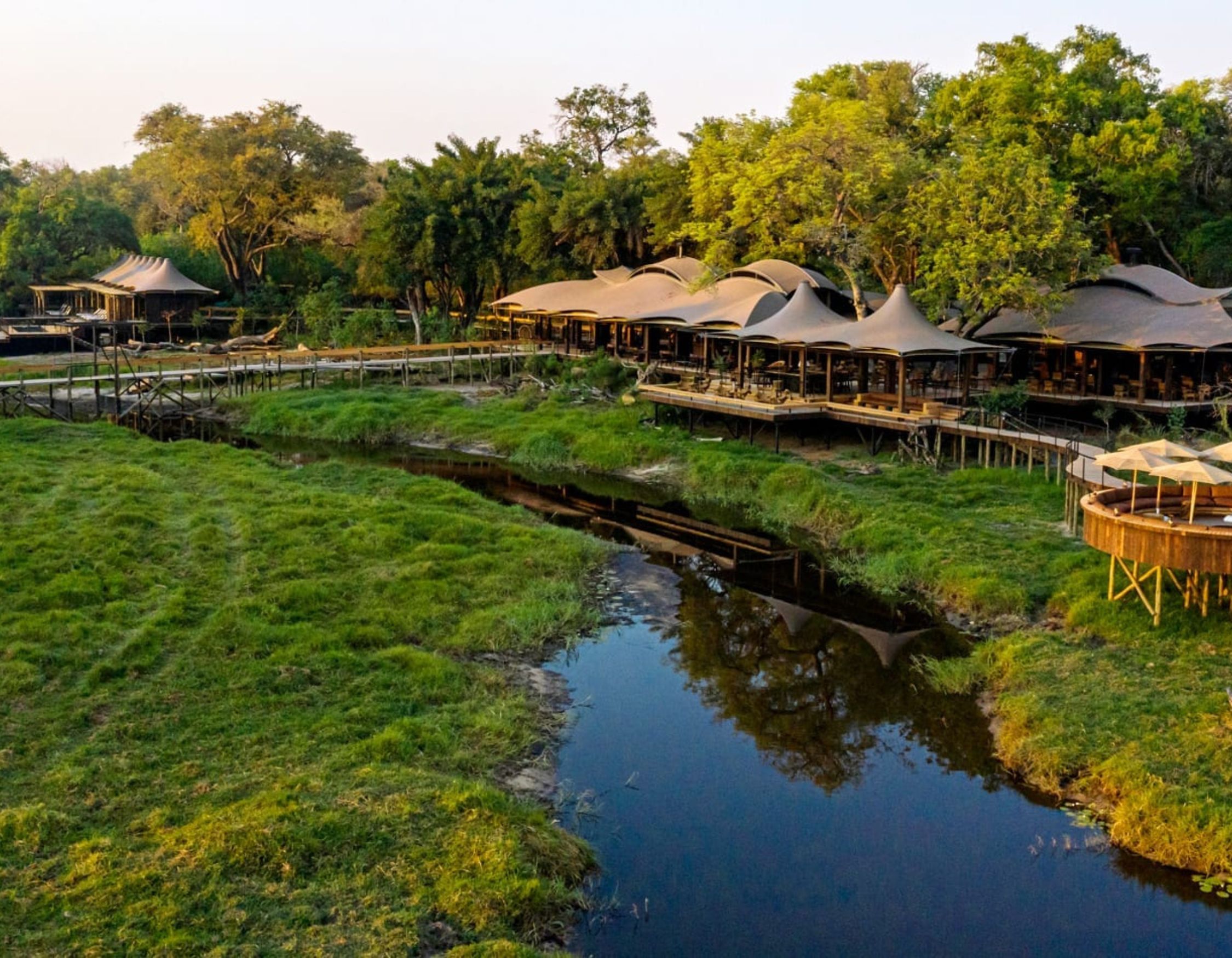 A scenic view of a safari lodge surrounded by lush greenery, featuring tent-like structures with thatched roofs. A tranquil stream flows nearby, reflecting the trees and open sky.