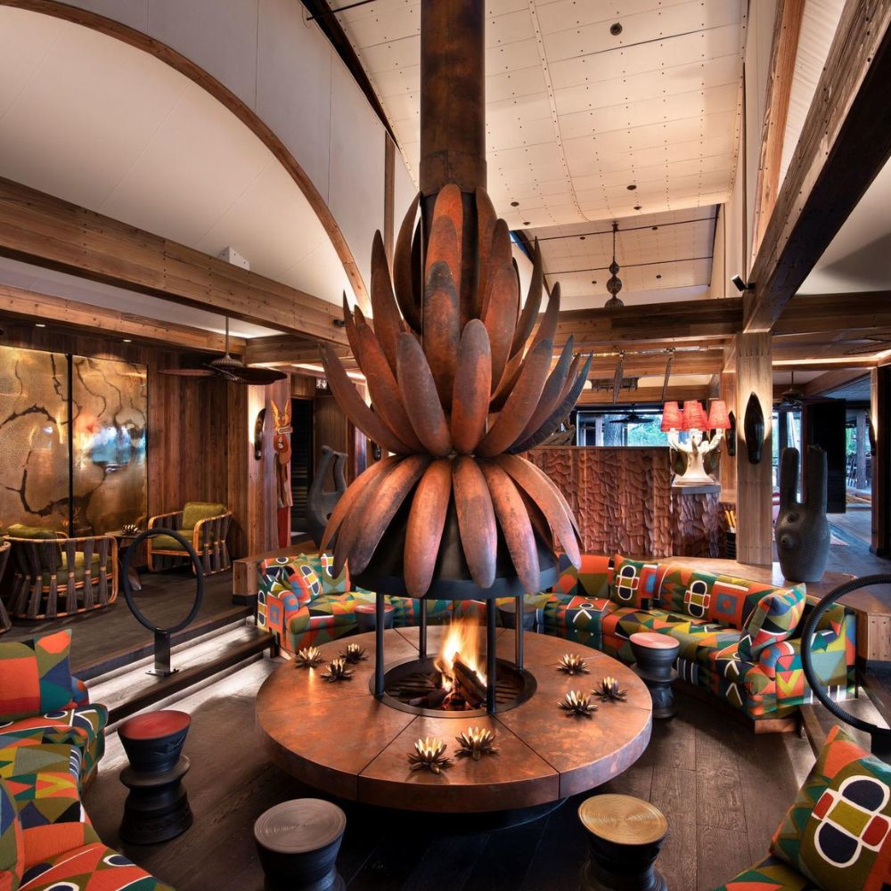 A cozy, modern lounge featuring a large circular fireplace with a striking floral design as the centerpiece. The seating area is adorned with vibrant, patterned cushions, and wooden accents create a warm atmosphere. Colorful artwork and sculptures are visible in the background.