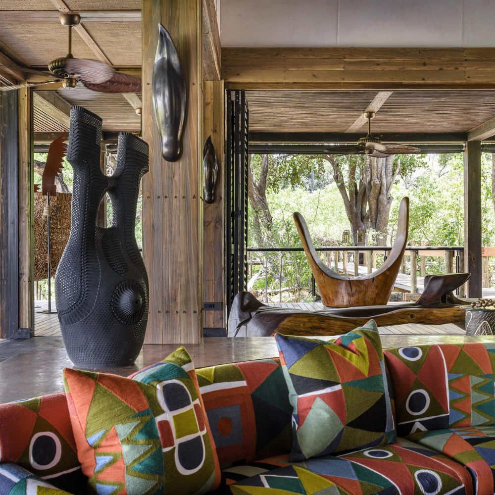 A stylish living space featuring a vibrant, patterned sofa with bold geometric designs. In the foreground, an intricately designed black sculpture stands beside a beautifully crafted wooden chair with a curved backrest, all set in a warm, wooden interior with large windows overlooking greenery.