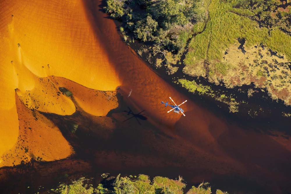 A blue helicopter hovers above a river with vibrant orange and brown sediment patterns, surrounded by lush green vegetation. The reflection of the helicopter creates a shadow on the water's surface.