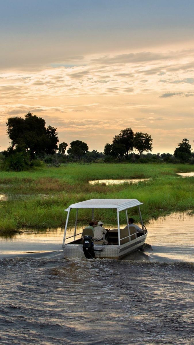 A boat with passengers navigates through a tranquil waterway, surrounded by lush greenery and trees, under a colorful sunset sky.