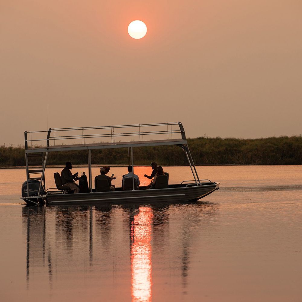 A group of four people on a boat silhouettes against a serene sunset, with the sun reflecting on the calm water.
