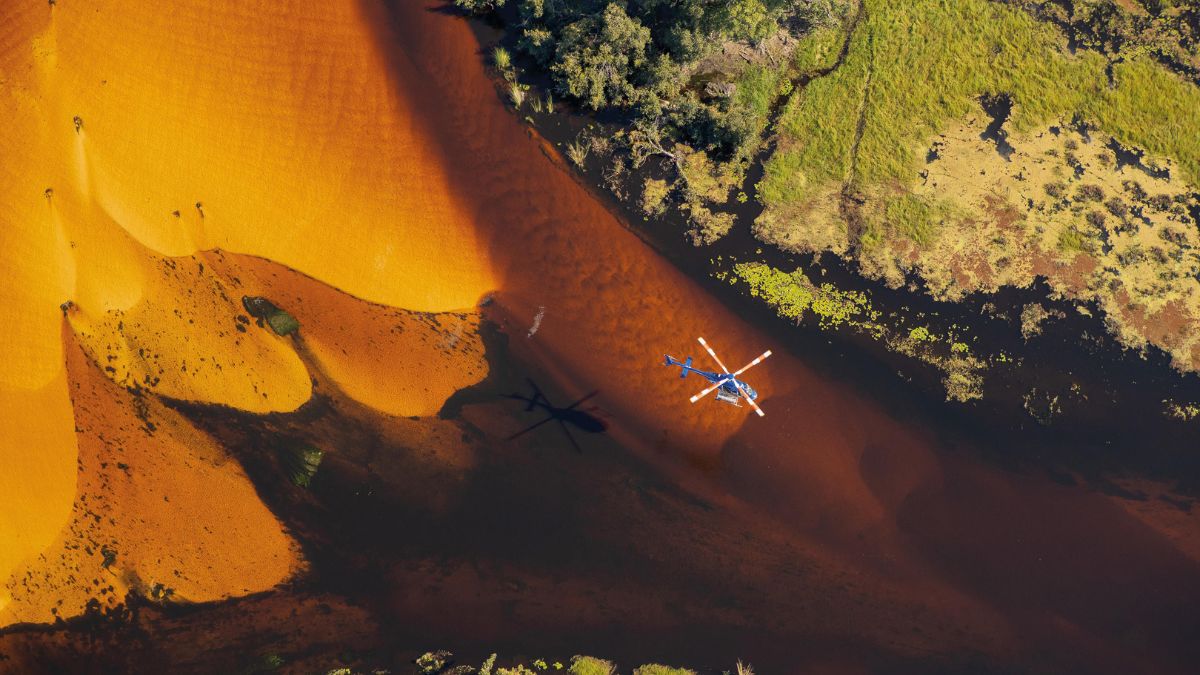 A blue helicopter hovers above a winding river surrounded by vivid orange sand dunes and lush green vegetation. The shadow of the helicopter stretches across the landscape below.