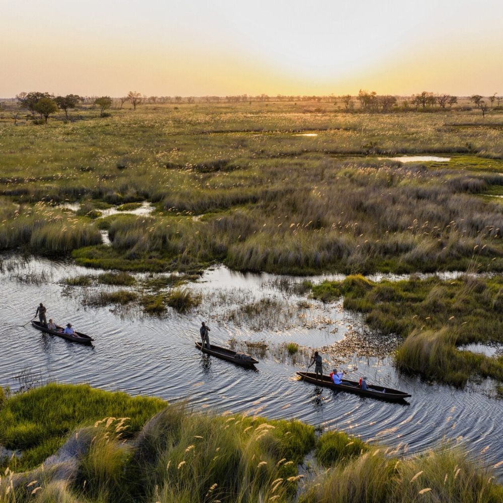Three wooden boats navigate through a winding river surrounded by lush green marshlands at sunset. The sky is painted in warm hues of orange and purple, casting a serene glow over the landscape.