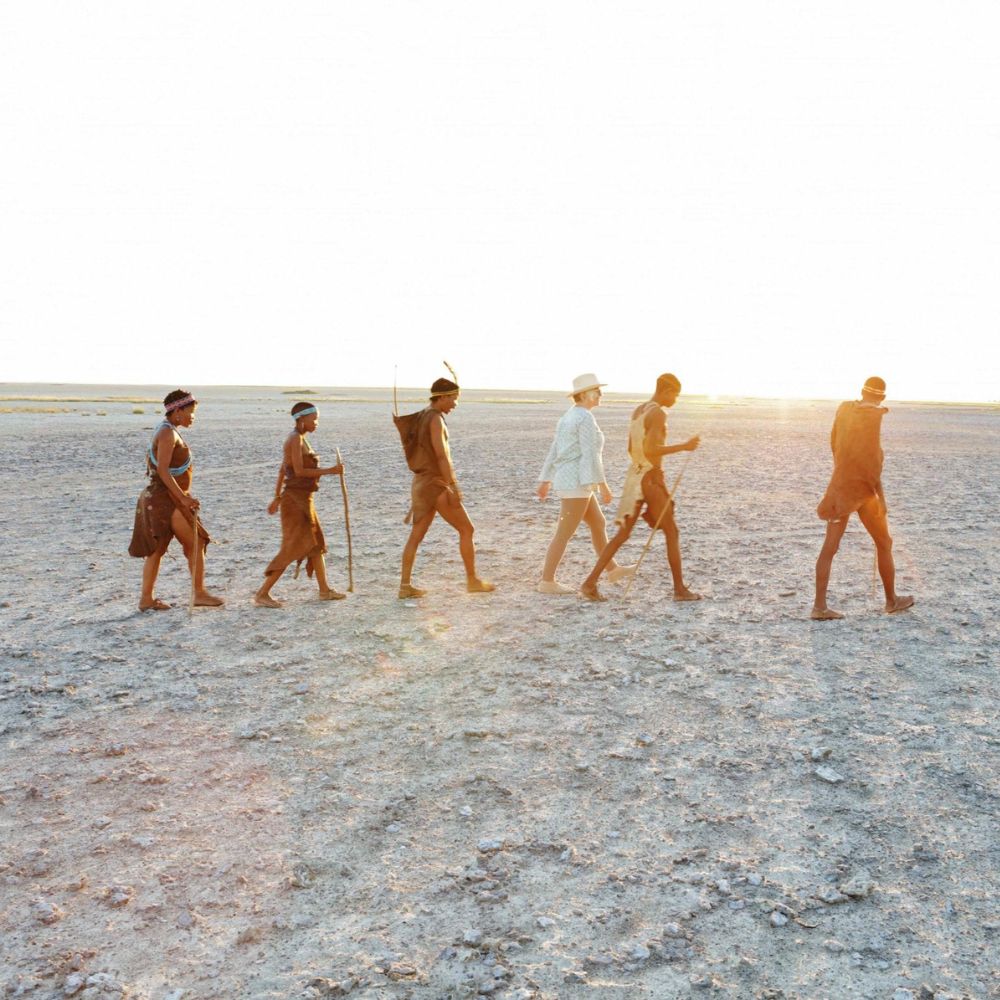 A group of people, including individuals wearing minimal clothing, walking in a line across a dry, desolate landscape with a bright sun setting in the background.