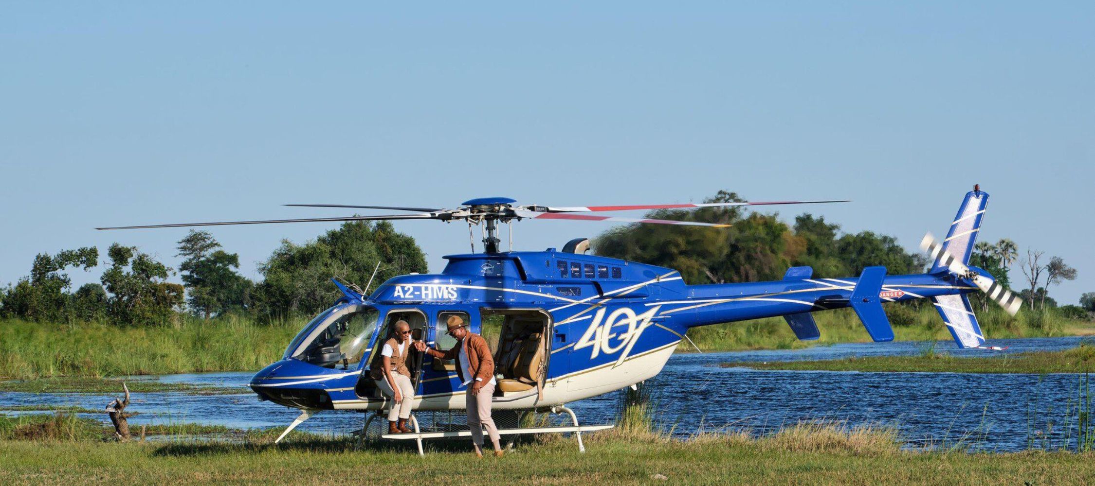 A blue helicopter is parked near a body of water, with two people preparing to board. Lush greenery surrounds the scene, indicating a scenic outdoor location.