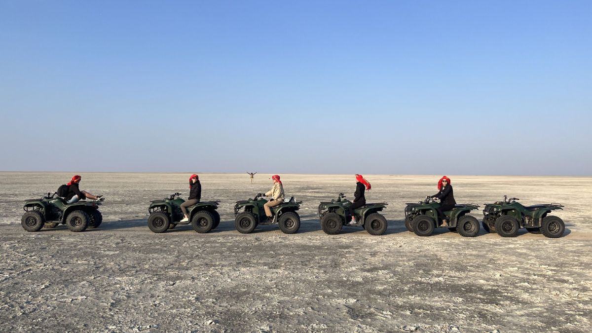 A group of six people wearing red headscarves are riding all-terrain vehicles on a vast, dry salt flat, with a clear blue sky above.