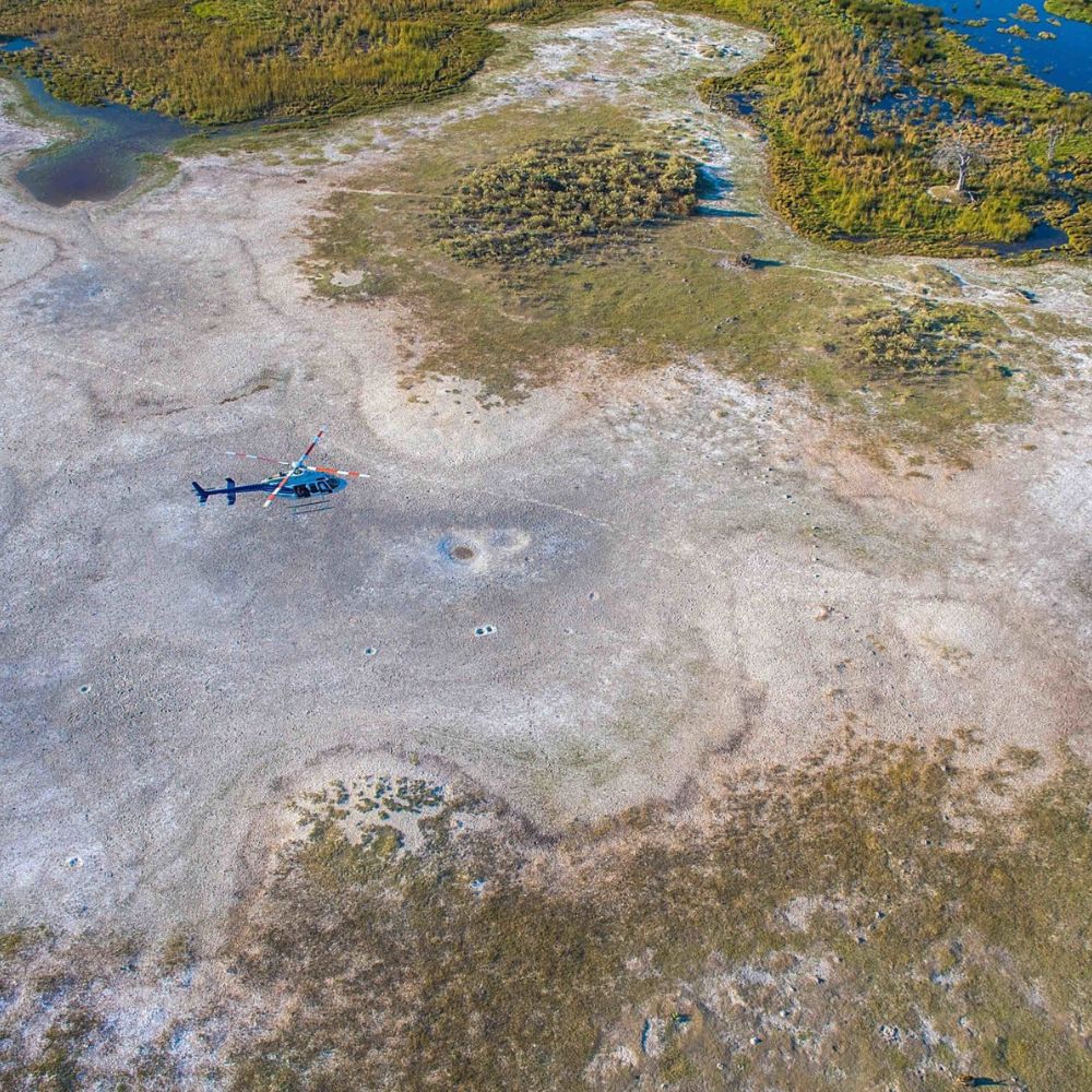A helicopter sits on a dry, grassy terrain surrounded by patches of water and greenery, viewed from above.