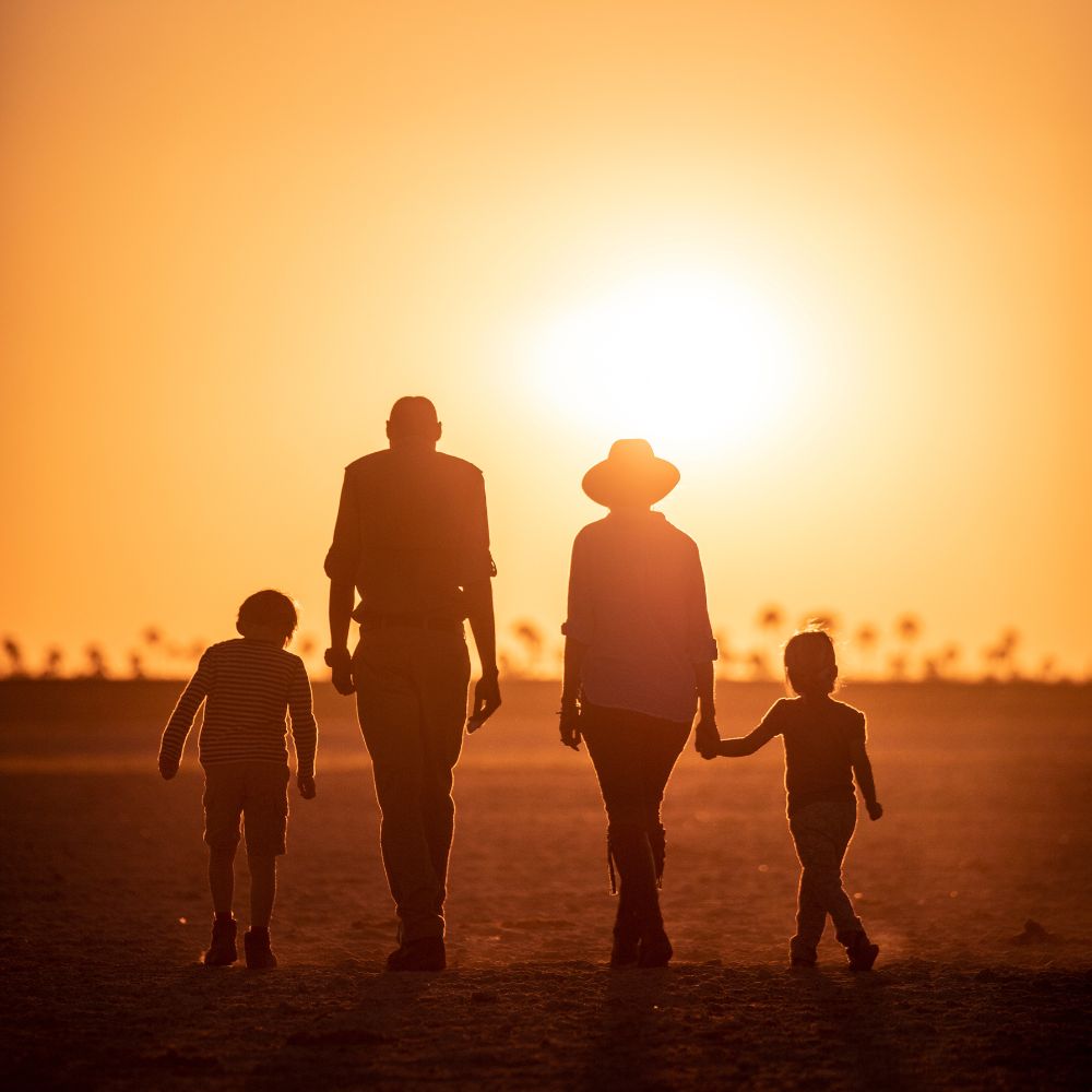 A family of four walks hand in hand along a sandy path during sunset, silhouetted against a glowing orange sun. Palm trees are faintly visible in the background.