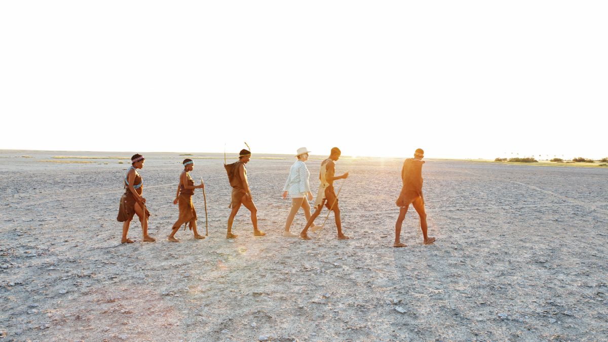 A group of six individuals walking in a line across a dry, open landscape during sunset. They are dressed in traditional attire, with some holding spears, and the sun casts a warm glow on the scene.
