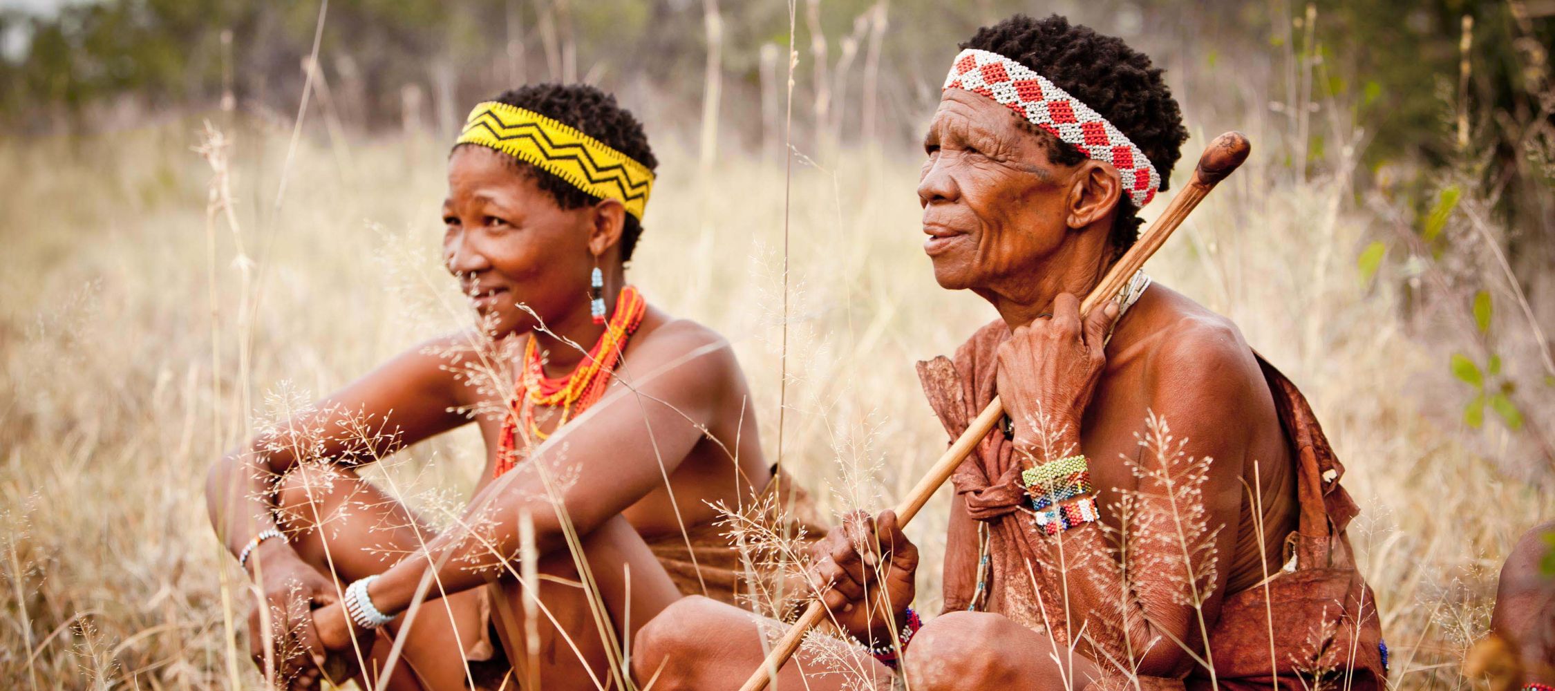 Two women in traditional attire sit on dry grass in a natural setting. The younger woman smiles and wears colorful beaded jewelry and a yellow headband, while the older woman, holding a stick, gazes thoughtfully into the distance, adorned with intricate jewelry and a patterned headband.