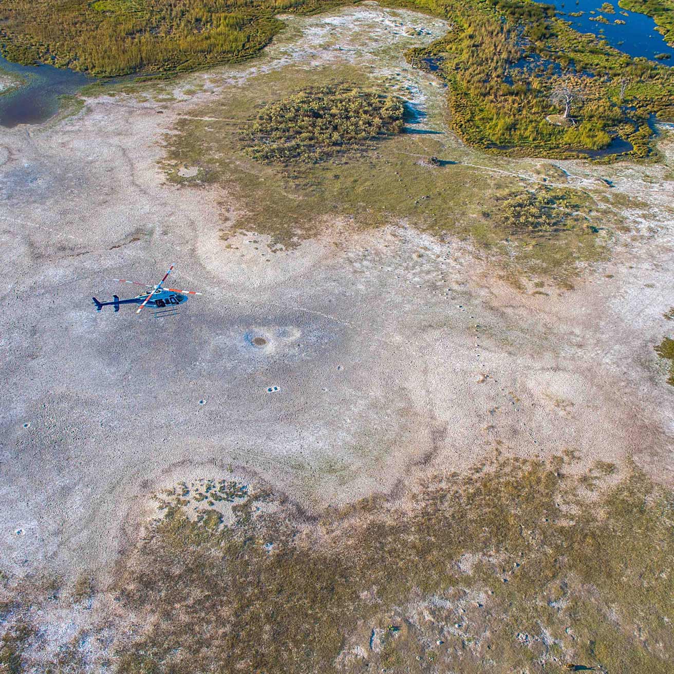 A helicopter is positioned on dry, cracked earth surrounded by patches of green vegetation and water in a vast, barren landscape.