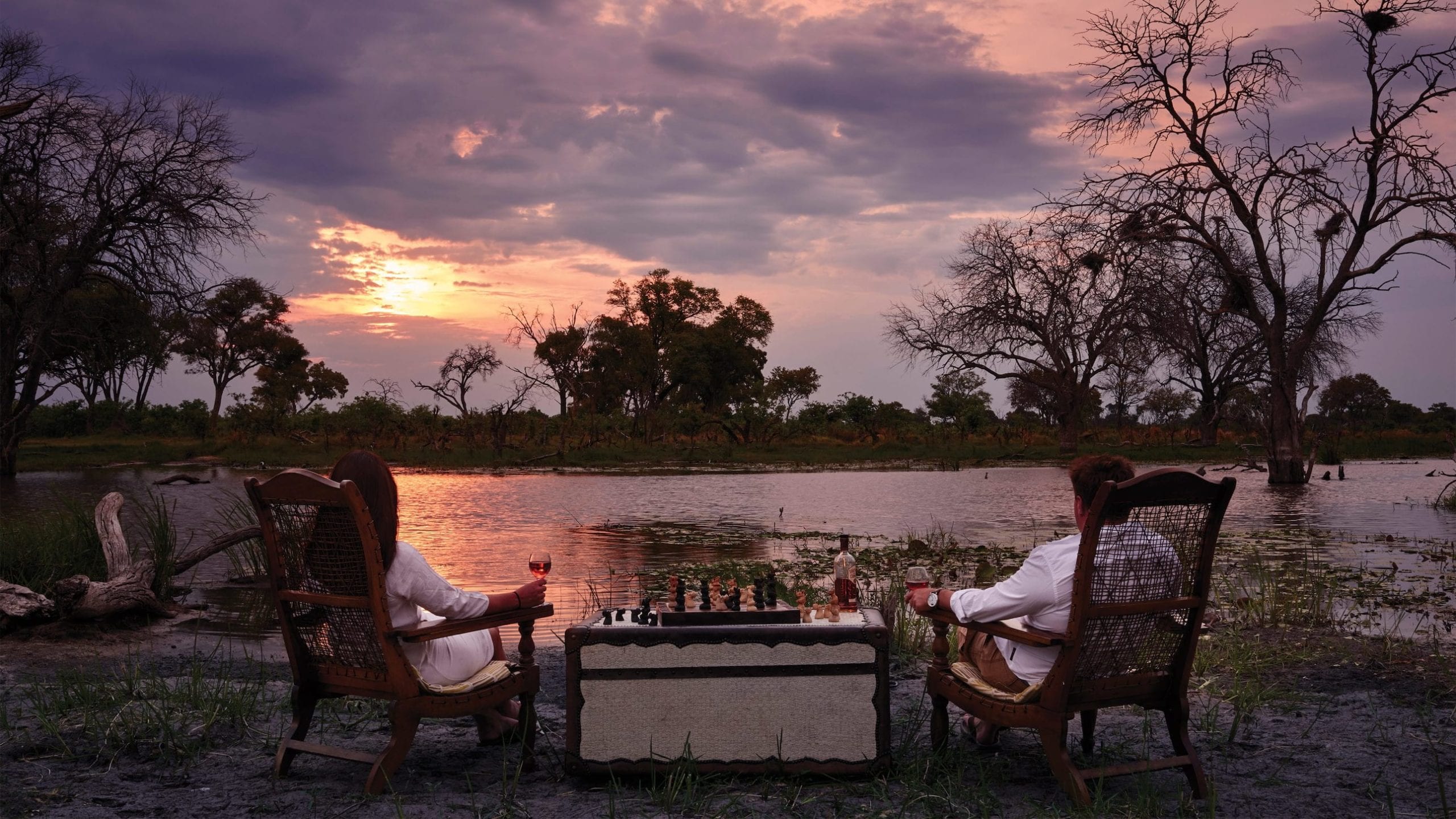 Two people sit in elegant chairs facing a serene landscape with a sunset over a body of water. They hold glasses of red wine, while a chessboard sits on a table between them. Silhouetted trees and foliage frame the tranquil scene.
