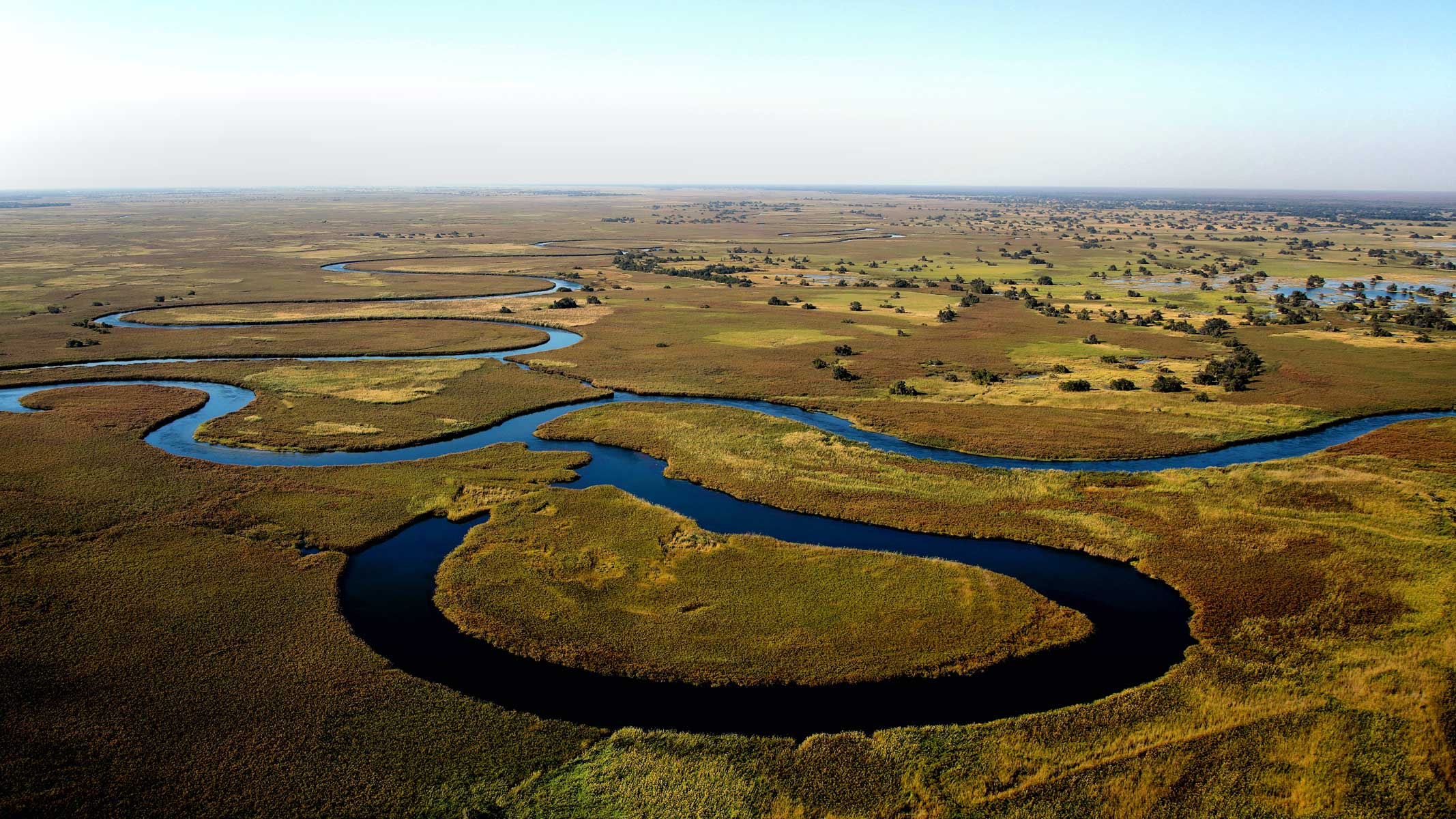 Meandering river flows through a lush, green landscape with patches of dry grass and scattered trees under a clear blue sky.