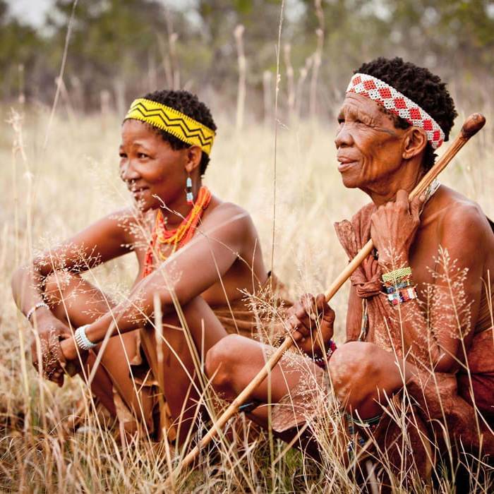 Two women sitting in tall grass, wearing traditional attire and beaded accessories. One holds a wooden staff and both have elaborate headbands. They appear engaged and thoughtful.