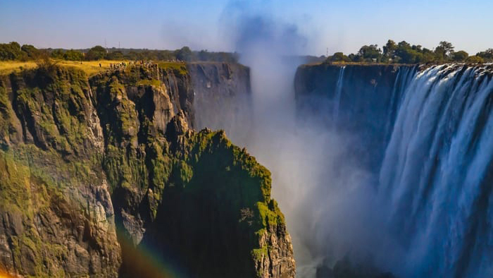 Victoria Falls cascades dramatically over steep cliffs, creating a misty atmosphere. Lush greenery surrounds the falls, with visitors visible at the edge. A rainbow forms in the mist under a clear blue sky.