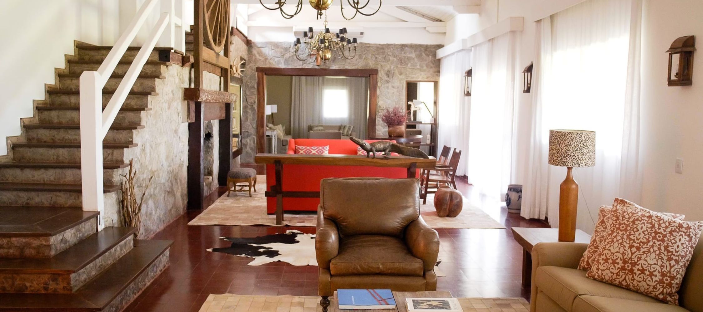 A spacious living area with a mix of modern and rustic decor. The room features a leather armchair, a cozy sofa, a coffee table, and a large wooden dining table. A staircase leads to the upper floor, and there are elegant light fixtures hanging from the ceiling. Natural light filters through large windows, illuminating the stone walls and wooden accents.